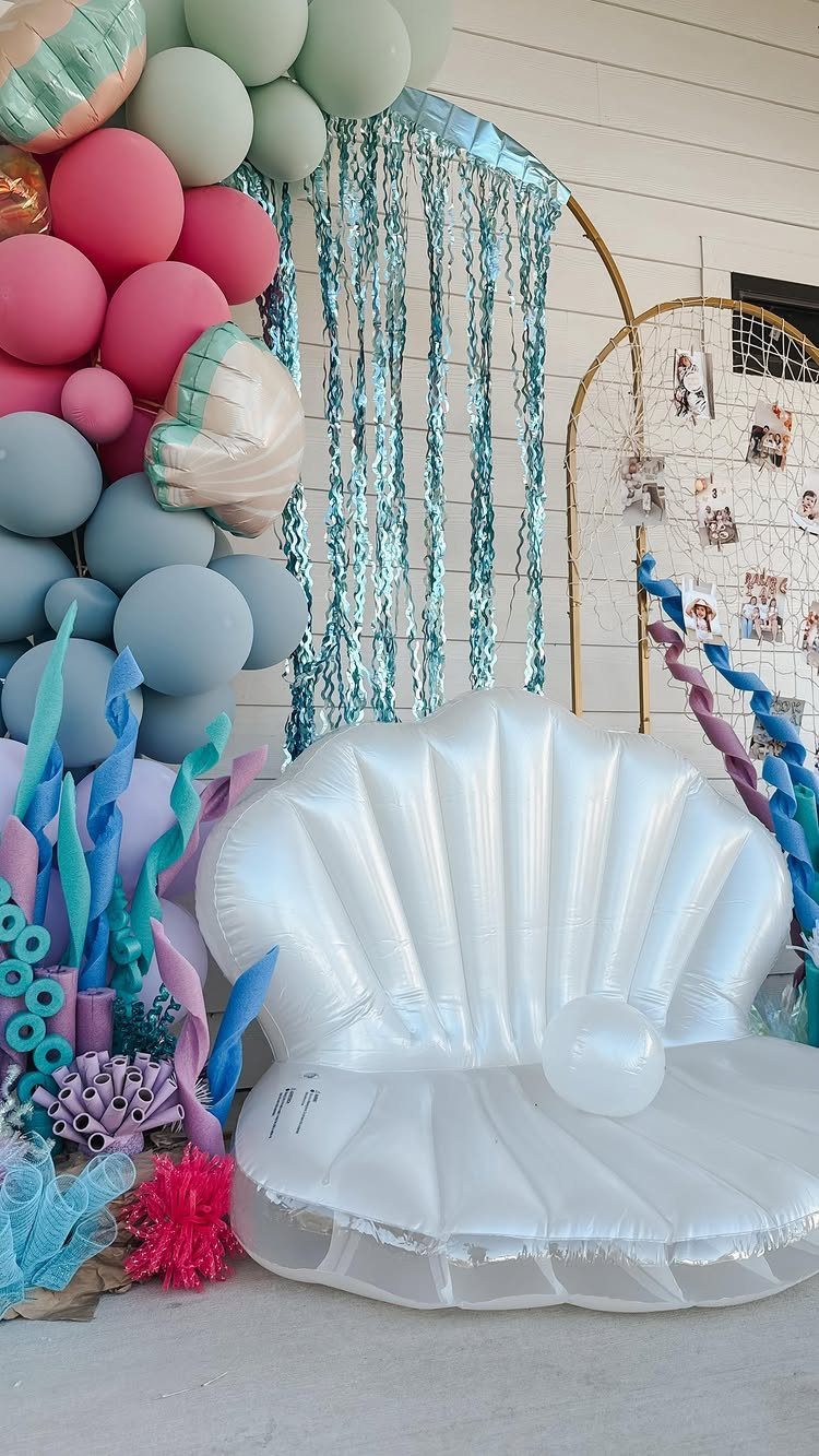 White inflatable seashell chair surrounded by blue pool noodle coral and colorful balloons.