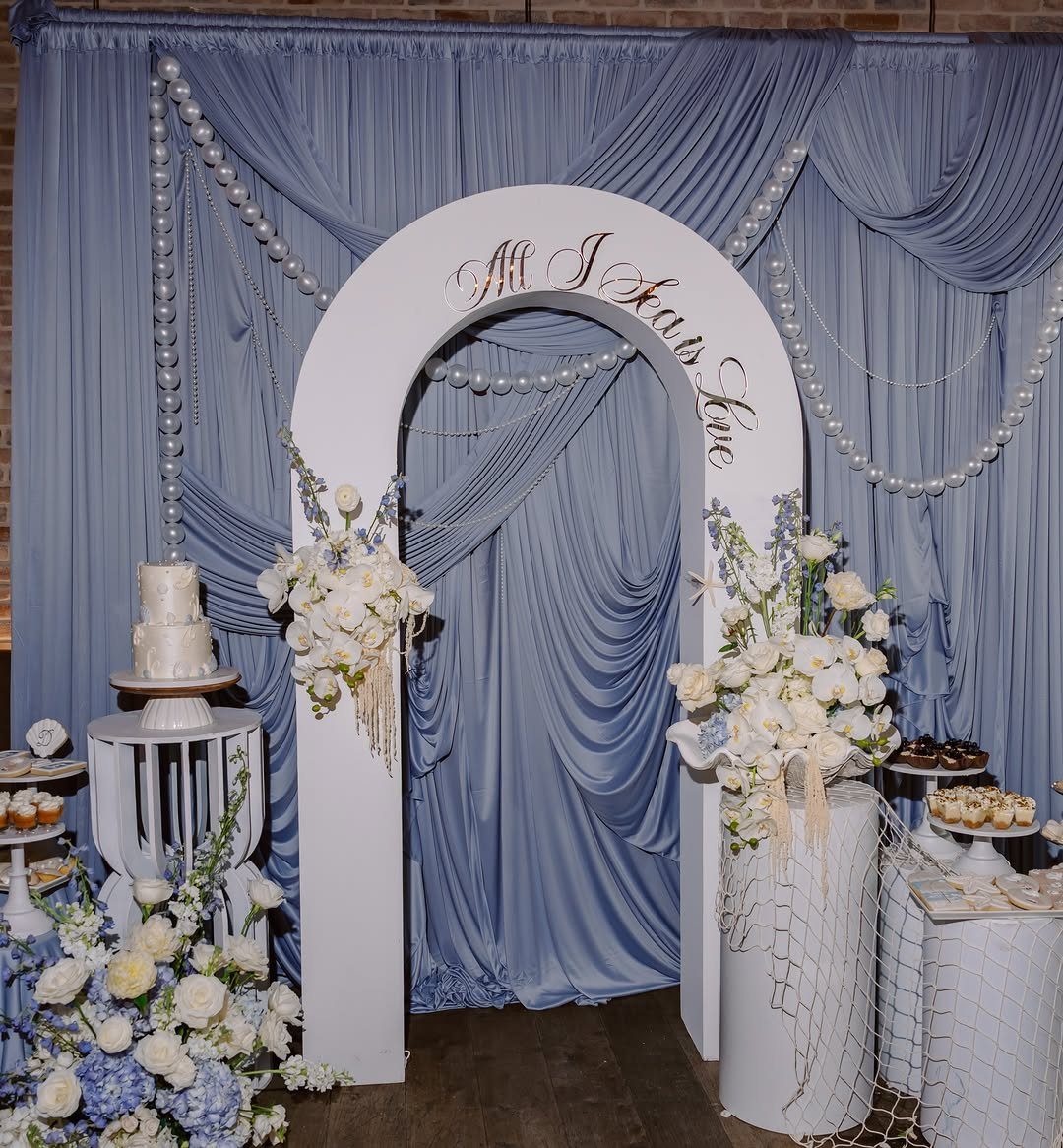 Blue fabric drapery backdrop with pearl garlands, white floral arrangements, and an arch.