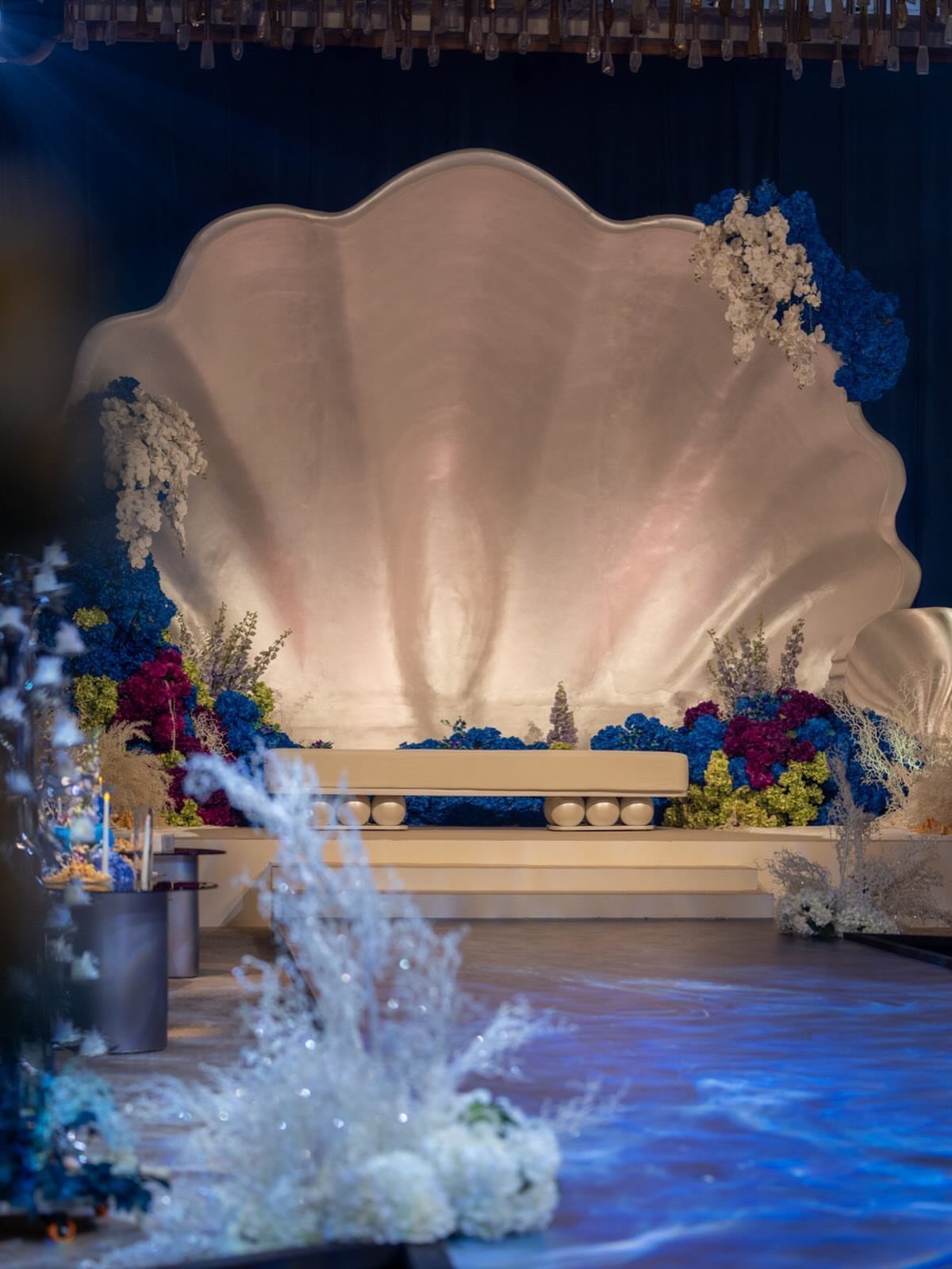 Giant iridescent seashell backdrop surrounded by blue and purple flowers and coral structures.