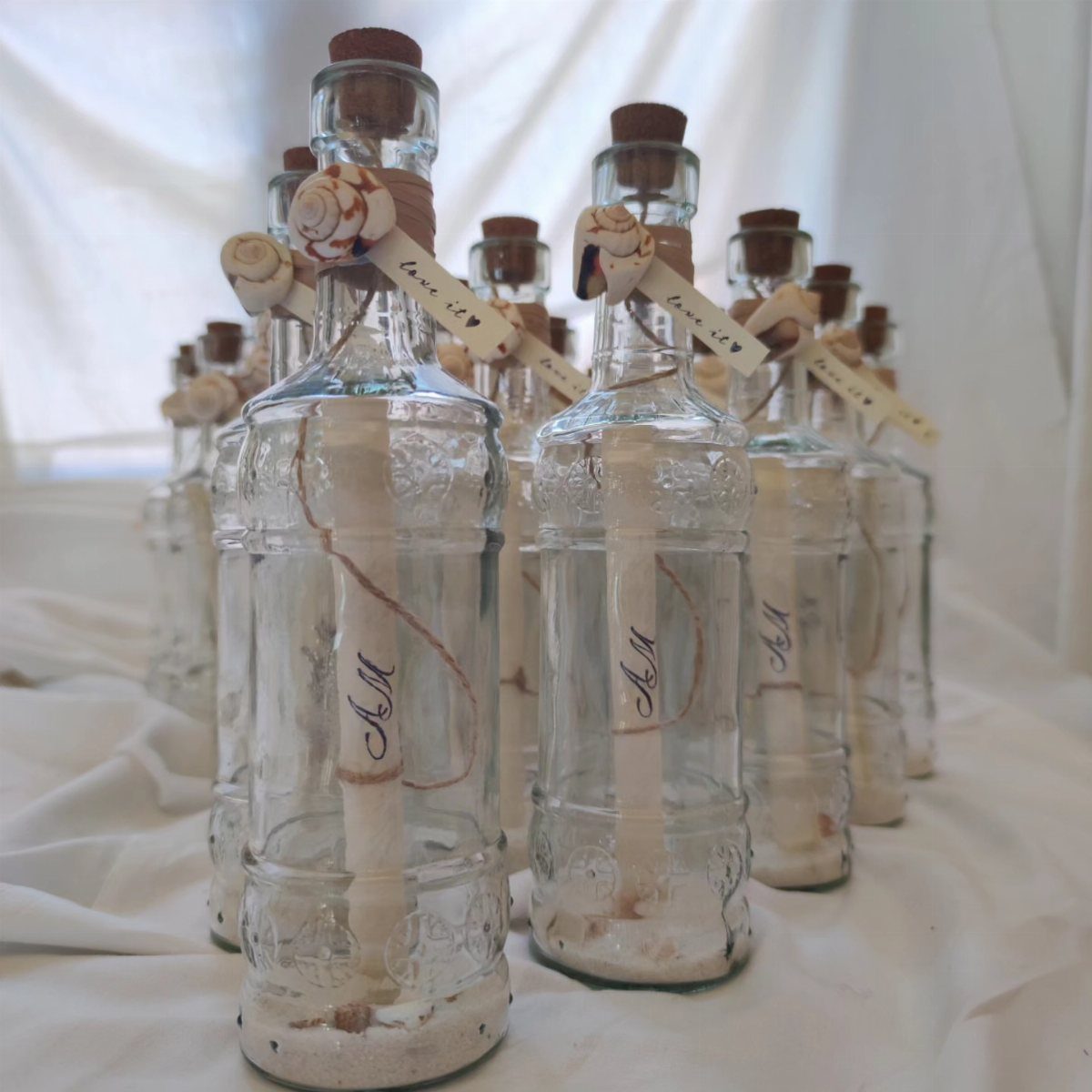 Rolled white paper invitations inside clear glass bottles decorated with small seashells and twine.