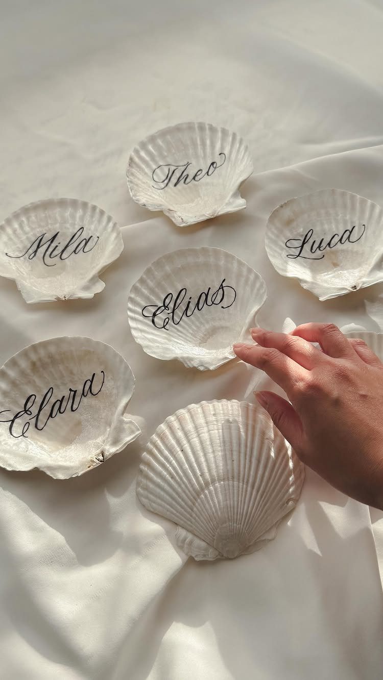 White scallop seashells with handwritten names used as place cards on white fabric.