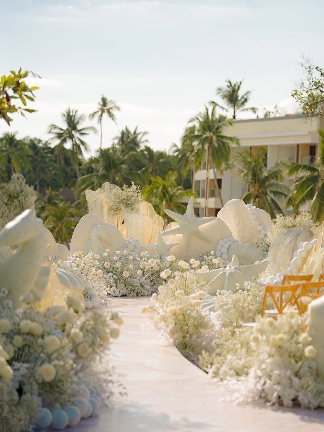 White sculptural seashells and starfish among white flowers lining an outdoor wedding aisle.