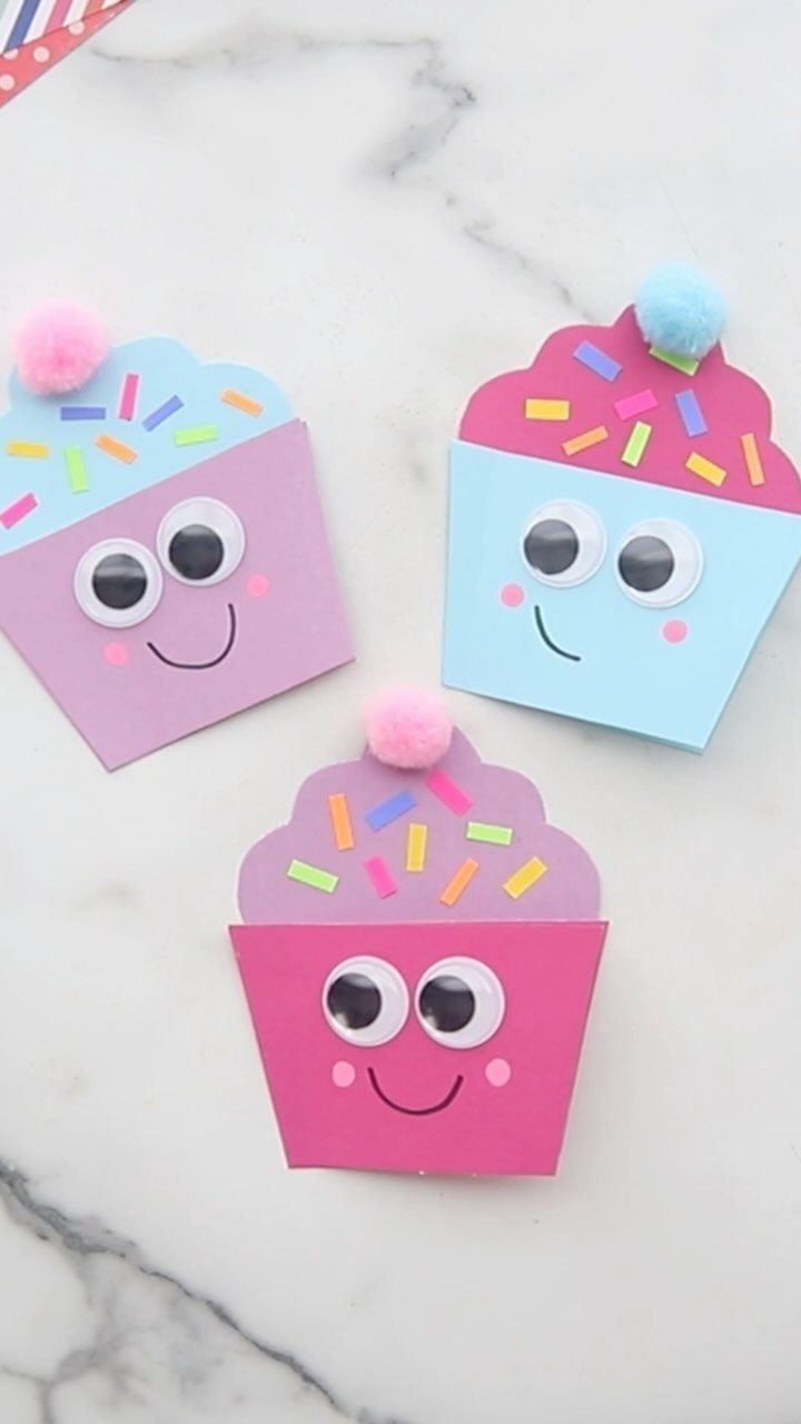 Three colorful handmade cupcake cards with googly eyes and pom-poms on marble surface.