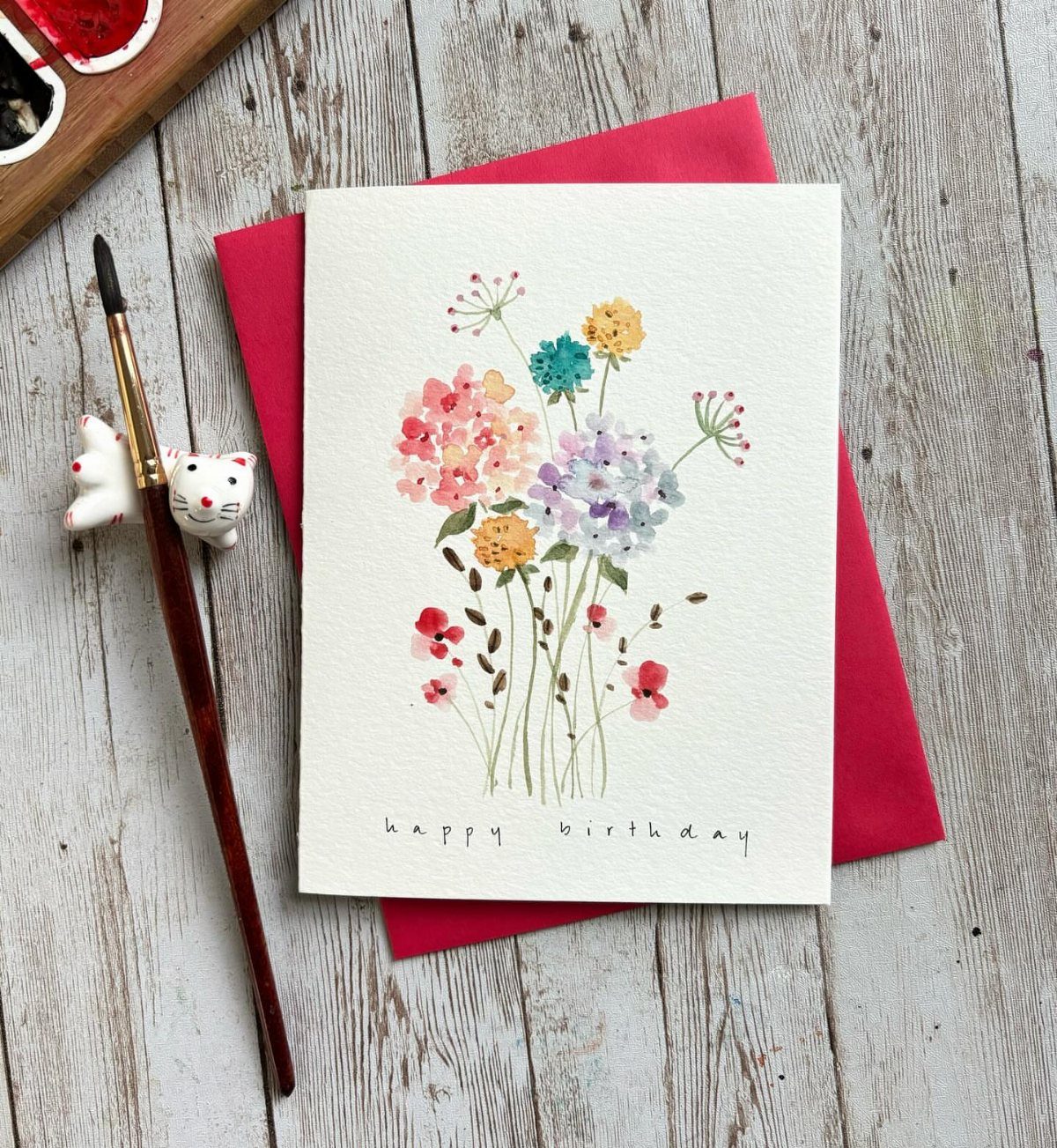 Watercolor birthday card with colorful flowers resting on a red envelope and wooden table