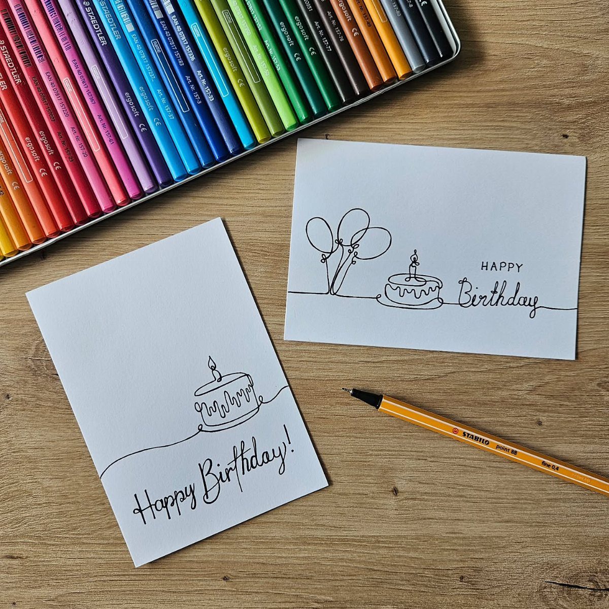 Two white birthday cards with minimalist black line art on a wooden table.