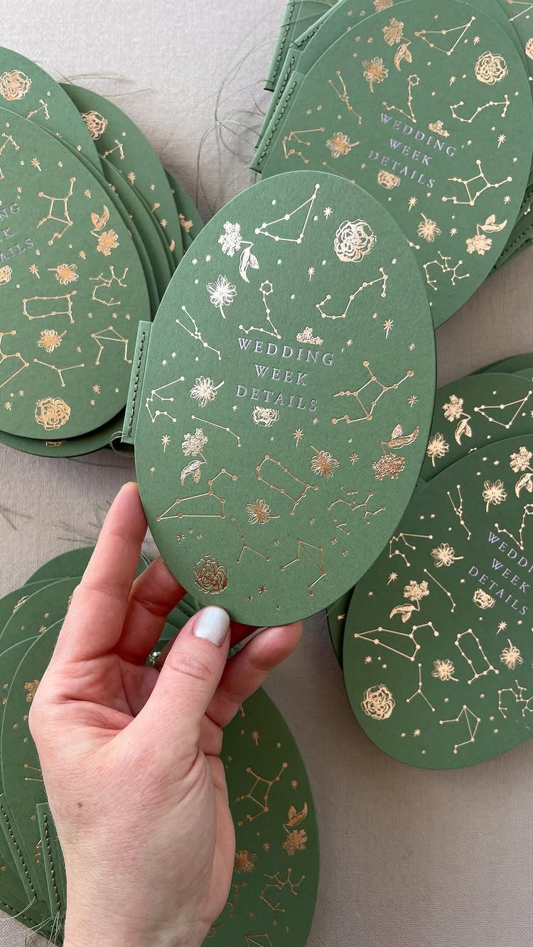 Green oval wedding week details cards with gold foil celestial constellation designs.