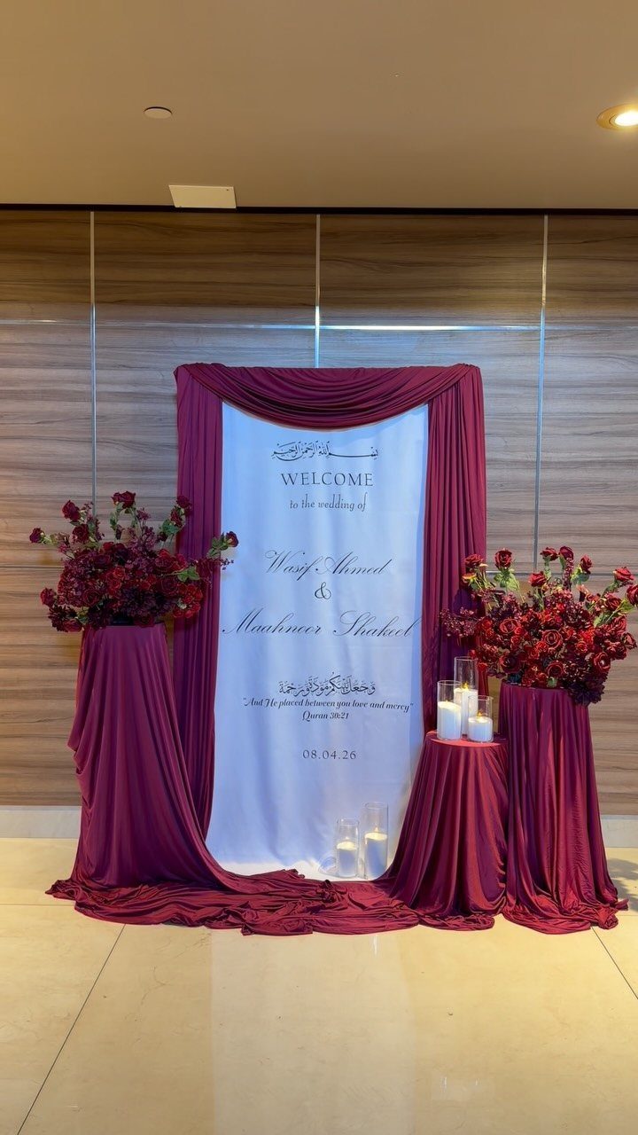 Burgundy draped fabric framing a white wedding welcome banner with red floral arrangements.