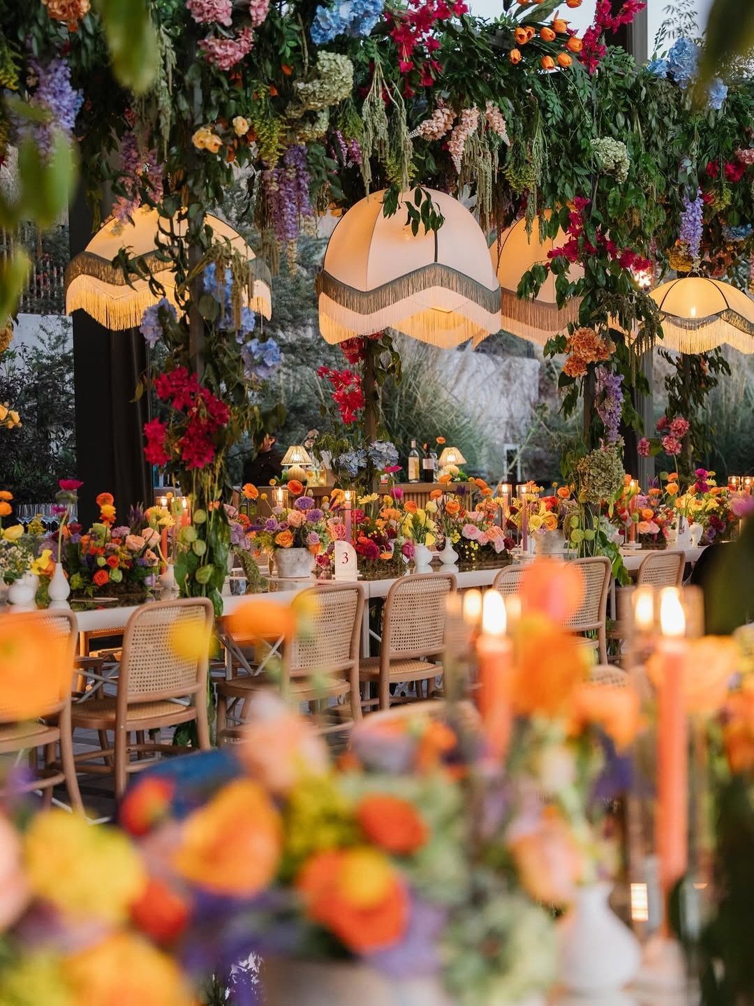 Decorated wedding reception table with colorful flowers, orange candles, and hanging dome lamps.